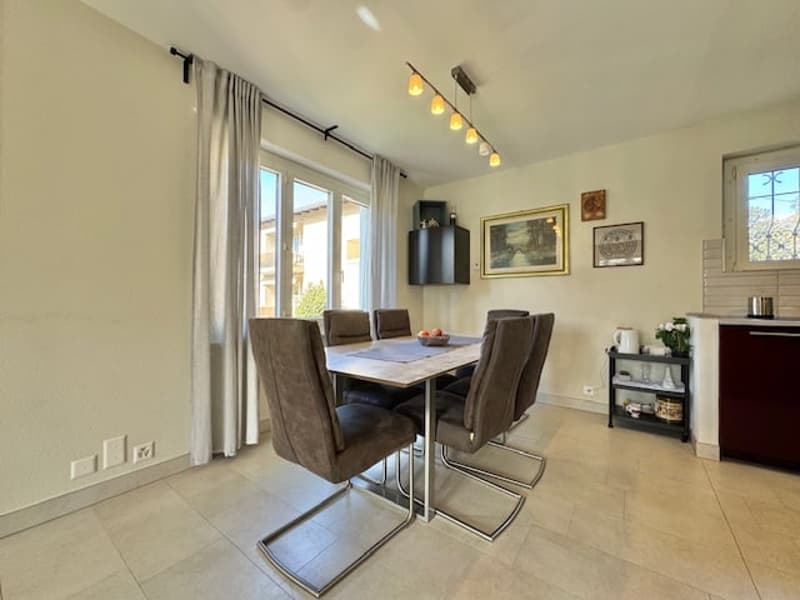 The image shows a spacious dining room with a large dining table and six chairs. The room has large windows, allowing natural light to enter. The walls are painted in a neutral color, and the floor is covered with tile. There are some decorative elements,