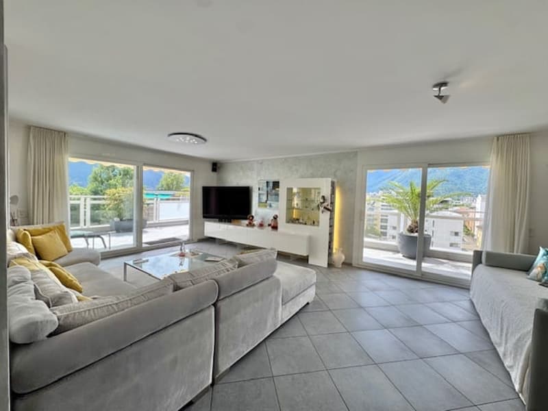 Spacious open-plan living room with large windows, gray sectional sofa, coffee table, and TV stand. The room has tiled flooring and access to a balcony or terrace.