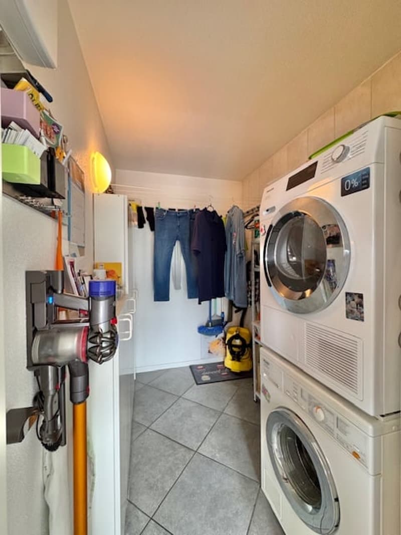 The image shows a small laundry room with a washing machine and a dryer stacked on top of each other. There are various cleaning supplies and tools stored on shelves, and clothes are hanging on a rack. The room has tiled flooring.