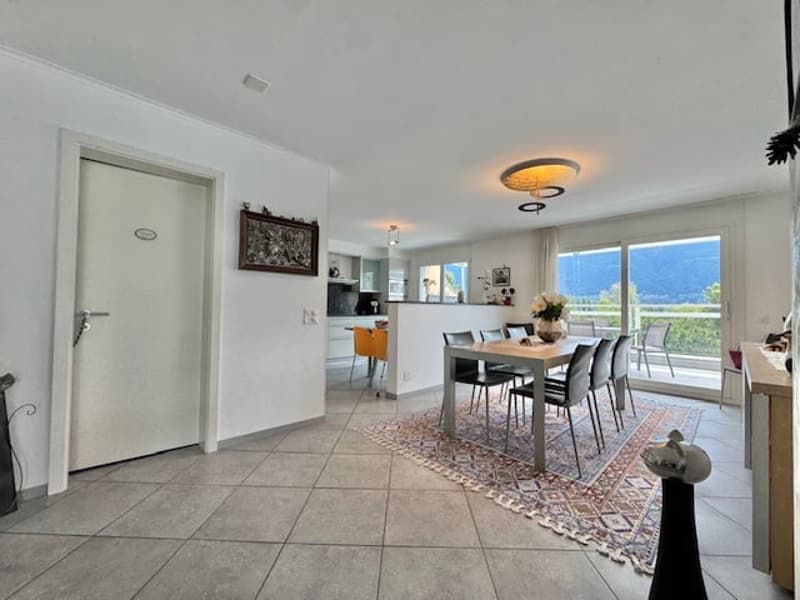 Open-concept living room and dining area, with large windows providing a view, tile flooring, dining table with chairs, and a decorative rug on the floor.