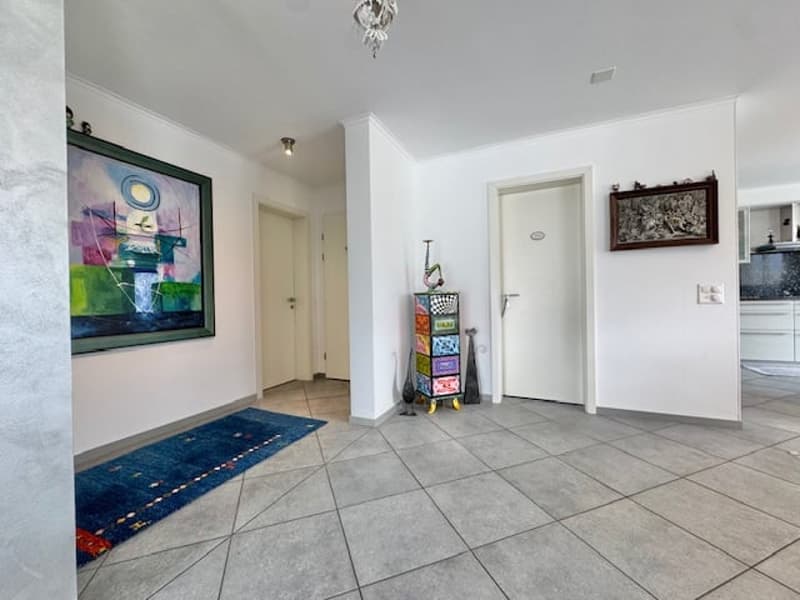 The image shows an apartment interior with a spacious entryway. The floor is covered in gray tile, and the walls are painted white. There is a large abstract painting on the wall, and a decorative shelf unit with various items displayed on it. The entrywa