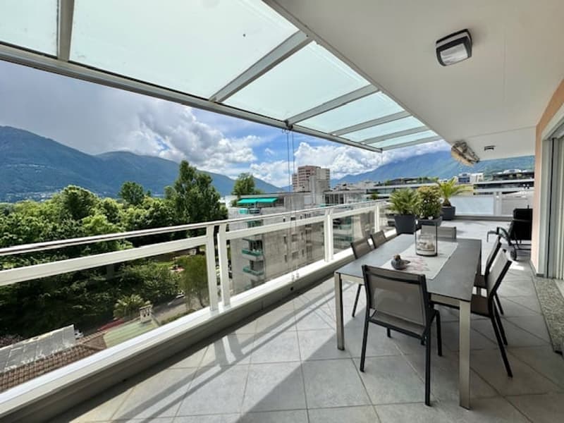 Spacious balcony with glass railing, outdoor dining table and chairs, overlooking a scenic mountain landscape with trees and buildings in the distance