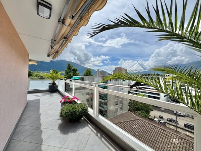 Spacious balcony with glass railing, overlooking a scenic view of mountains and buildings. The balcony is furnished with potted plants and has an open, airy feel.