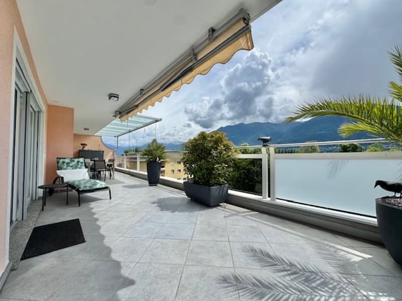 Spacious balcony with tiled flooring, retractable awning, potted plants, and outdoor furniture overlooking a scenic mountain landscape