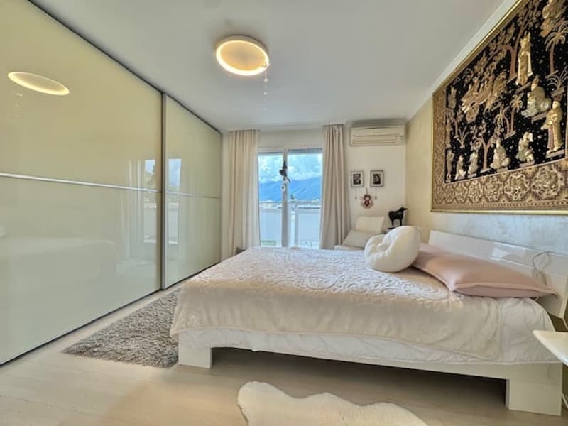 Spacious bedroom with large windows, sliding glass doors leading to a balcony, white bed frame and bedding, built-in closet, and a decorative wall hanging.