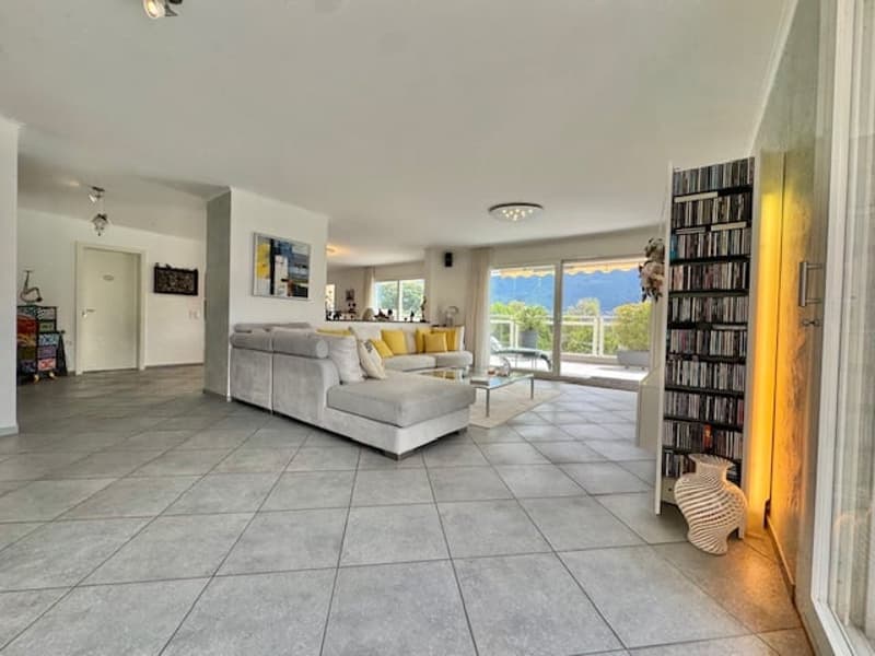 Spacious open-plan living room with large windows, grey tile flooring, grey sofa, yellow accent chair, bookshelf, and access to a balcony or terrace with a view