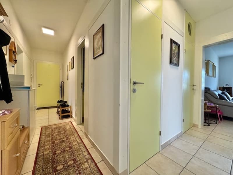 The image shows a hallway with a tiled floor, white walls, and several doors leading to other rooms. There is a wooden cabinet or dresser along the wall and some framed artwork hanging on the walls.