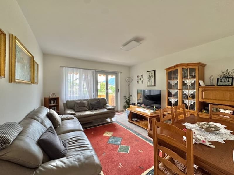Spacious living room with hardwood floors, gray sofas, wooden dining table and chairs, display cabinet, and large windows providing natural light.