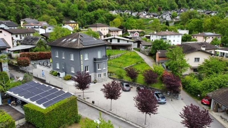 The image shows an aerial view of a residential area with a mix of multi-story buildings and houses surrounded by lush greenery. The buildings have solar panels installed on their roofs, and there are trees and landscaping throughout the area. The streets