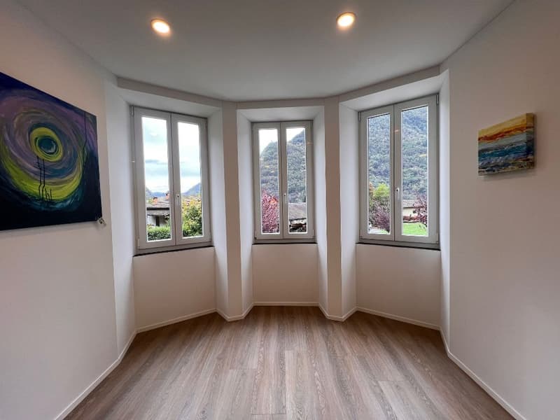 Spacious room with large windows offering a scenic view of the surrounding landscape. The room has a hardwood floor and white walls, with two paintings hanging on the walls. The room is well-lit with recessed lighting.