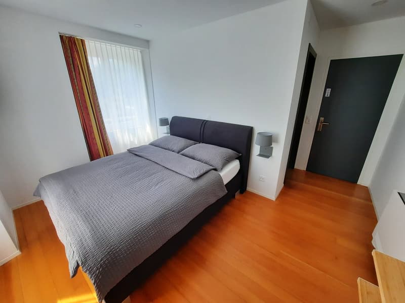 Spacious bedroom with hardwood flooring, white walls, large windows, a king-size bed with a gray comforter, and a balcony or terrace visible through the window.