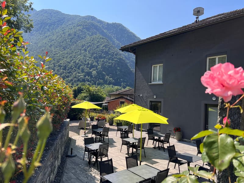 Outdoor seating area with yellow umbrellas, surrounded by lush green mountains and vegetation. The building has a dark exterior with windows and appears to be a restaurant or cafe.