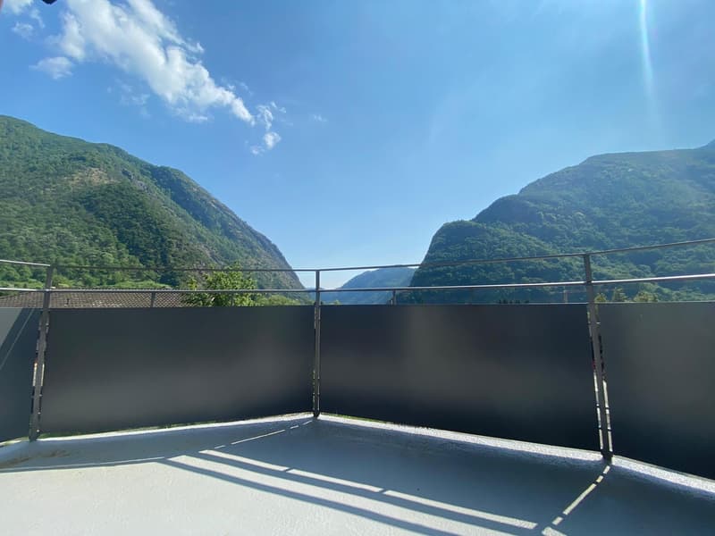 Outdoor balcony or terrace with a panoramic view of the surrounding mountains and lake. The balcony has a black railing and a concrete floor, and offers a scenic vista of the lush, green mountainous landscape.