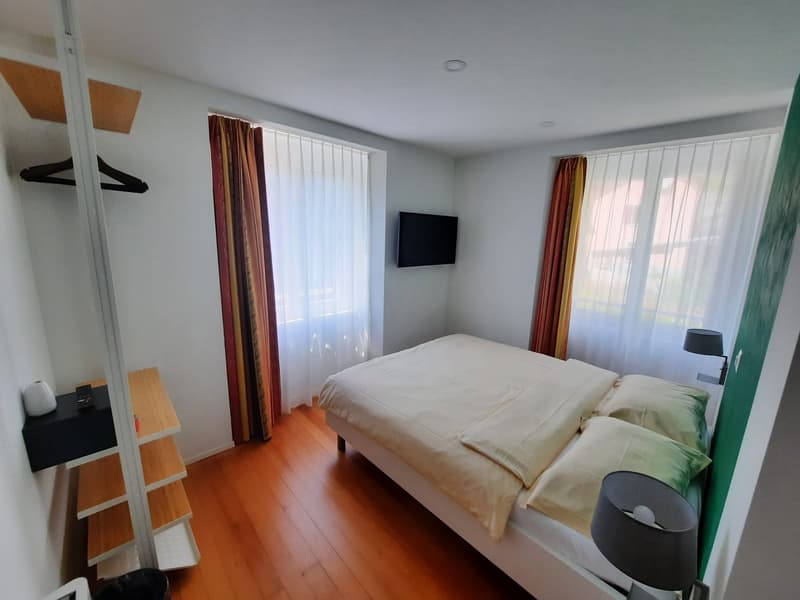 Spacious bedroom with hardwood flooring, white walls, large windows, a king-size bed, and curtains in warm colors. There is a TV mounted on the wall and a small side table with a lamp.