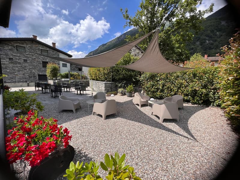 The image shows an outdoor seating area with a gravel floor, surrounded by lush greenery and mountains in the background. There are several white lounge chairs and tables set up under large canopy-like shade structures. The area appears to be part of a la
