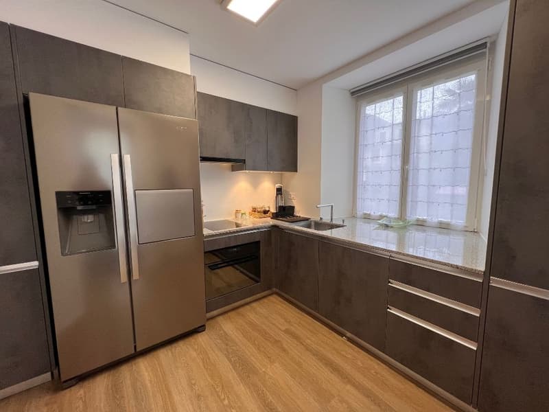 Modern kitchen with gray cabinets, stainless steel appliances including a refrigerator and dishwasher, and large windows providing natural light.