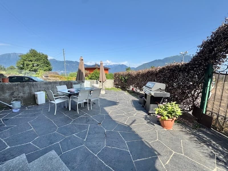 Outdoor patio with stone flooring, patio furniture, umbrellas, and a barbecue grill. The patio is surrounded by greenery and has a view of the mountains in the background.