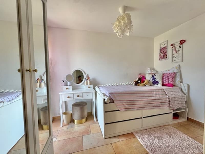 Spacious bedroom with white furniture, including a dresser with a mirror, a bed with drawers underneath, and a chandelier-style light fixture. The room has tile flooring and appears to have a balcony or terrace visible through the window.