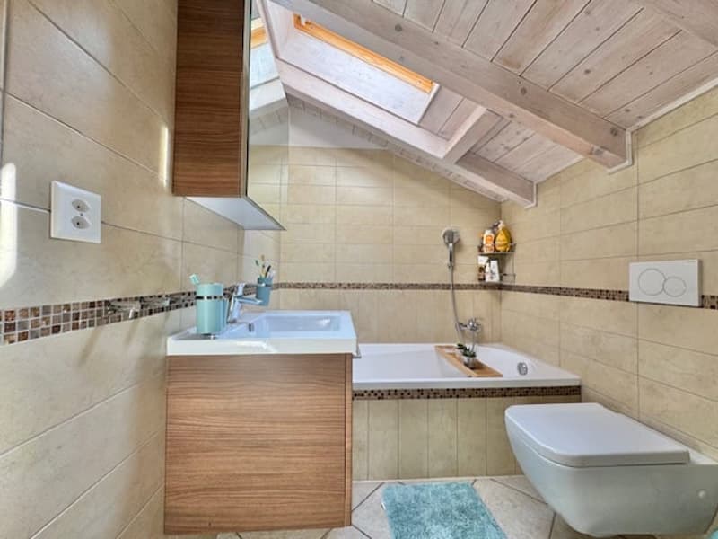 The image shows a bathroom with a wooden ceiling, tiled walls, a vanity with a sink, a toilet, and a bathtub.