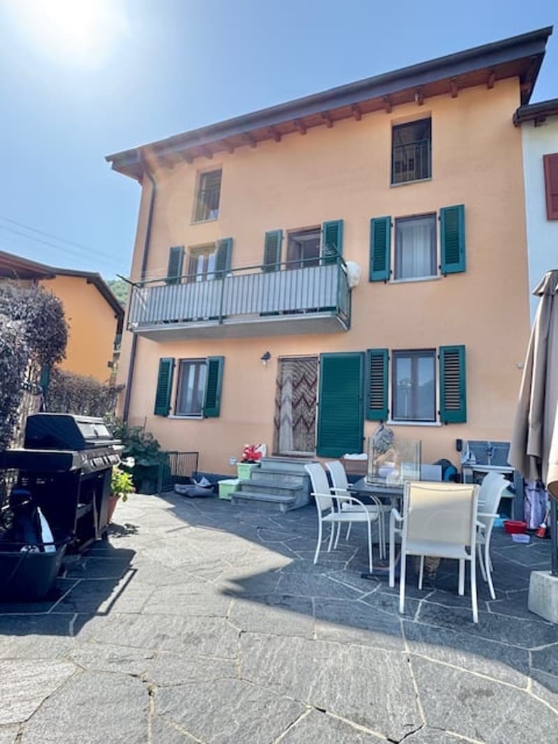 2-story house with yellow exterior, green shutters, balcony, patio with outdoor furniture, and stone paving in the front yard.