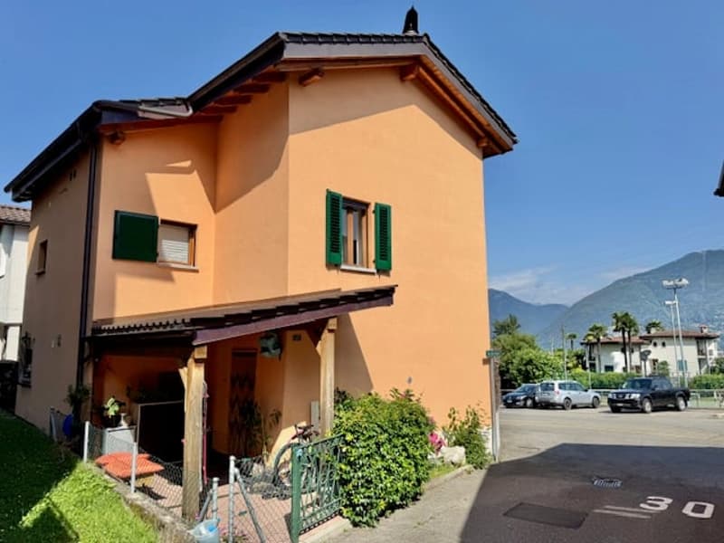 Two-story house with orange exterior walls, green shutters, and a tiled roof. The house has a small garden area in front and is located near a parking lot.