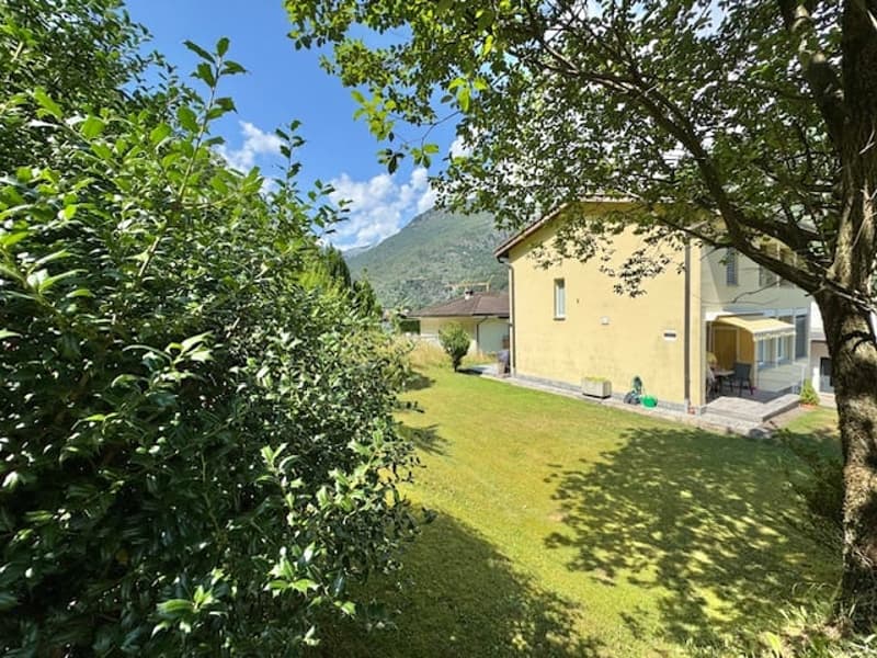Two-story yellow house with a balcony or terrace, surrounded by lush green trees and a well-maintained lawn.