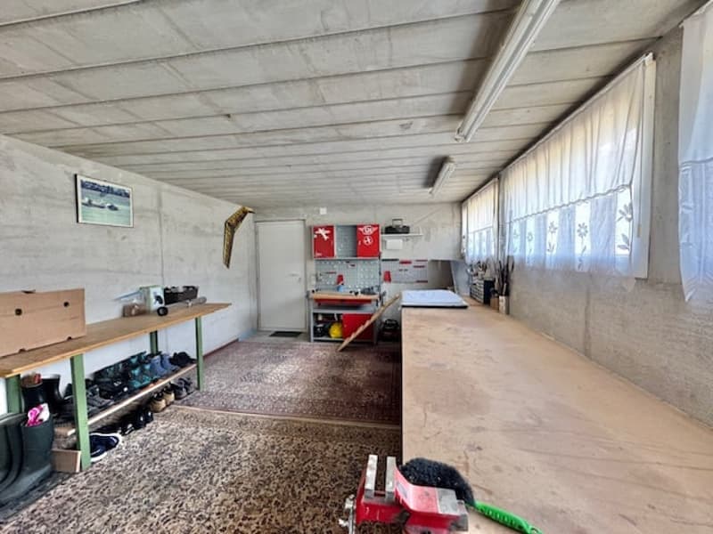 The image shows a garage or storage space with a concrete floor, shelves and storage units along the walls, and various tools and equipment scattered throughout the room.