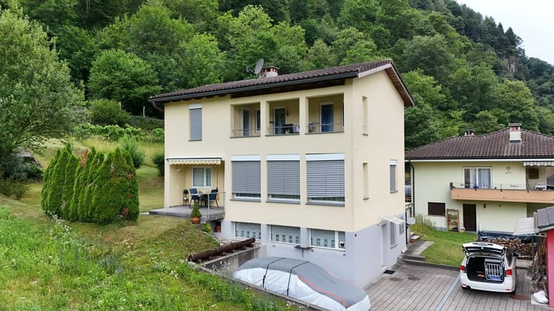 Two-story house with a yellow exterior, balconies, and a paved parking area. The house is surrounded by lush greenery and trees, creating a scenic, rural setting.