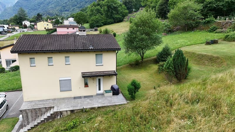 2-story house with yellow exterior, tiled roof, surrounded by lush green landscape with trees, shrubs, and a grassy area. The house has a balcony or terrace visible.