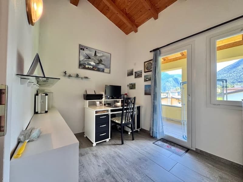 This image shows a room with a wooden ceiling, white walls, and a sliding glass door leading to a balcony or terrace. The room has a desk with a computer setup, and there are various decorative items and artwork on the walls.