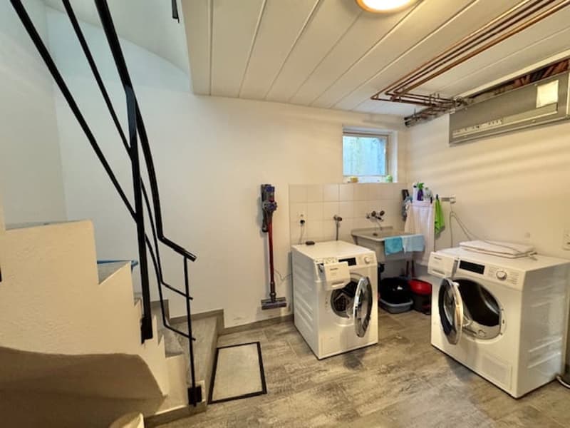The image shows a laundry room with a washer, dryer, and other appliances. The room has a concrete floor and white walls, and there is a window providing natural light.