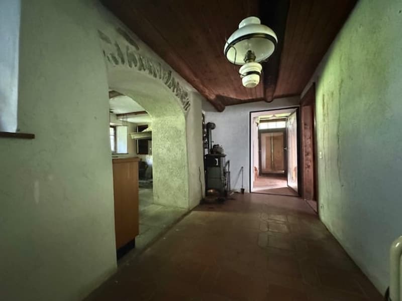 The image shows a long, dimly lit hallway with green walls and an arched entryway. The hallway has wooden floors and a ceiling with hanging light fixtures. There appears to be a doorway or entrance at the end of the hallway.