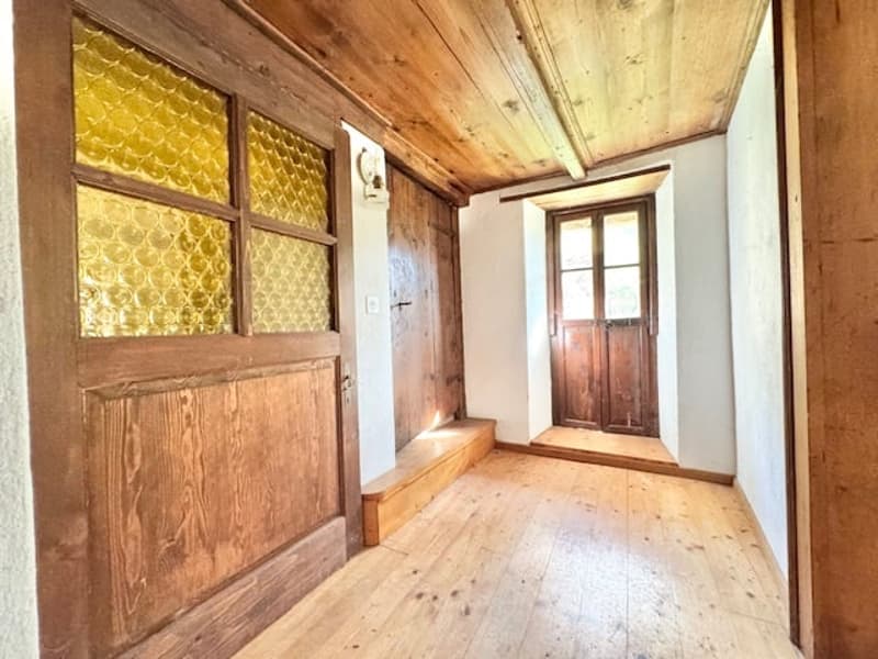 The image shows a hallway with wooden walls, wooden ceiling, and wooden floor. The hallway has a wooden door and a window with a decorative pattern on the glass.
