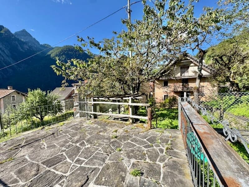 Two-story stone house with a wooden balcony or terrace, surrounded by blooming trees and mountains in the background.