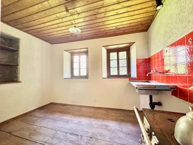 This image shows a room with a wooden ceiling, wooden floor, and white walls. The room has two windows and a red tiled wall with a sink or counter. The room appears to be a utility or laundry room based on the simple furnishings and layout.