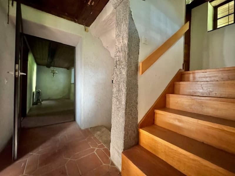 The image shows the interior of a building with a hallway and a staircase. The walls are made of stone and the floor is covered with terracotta tiles. The staircase is made of wood and has a simple, rustic design. The overall atmosphere appears to be that