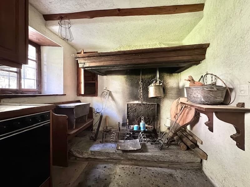 Old-fashioned kitchen with a large fireplace, wooden beams, and various antique cooking and household items such as pots, pans, and tools.