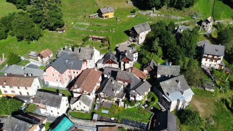 Aerial view of a small village nestled in a green, hilly landscape with houses, buildings, and gardens visible.
