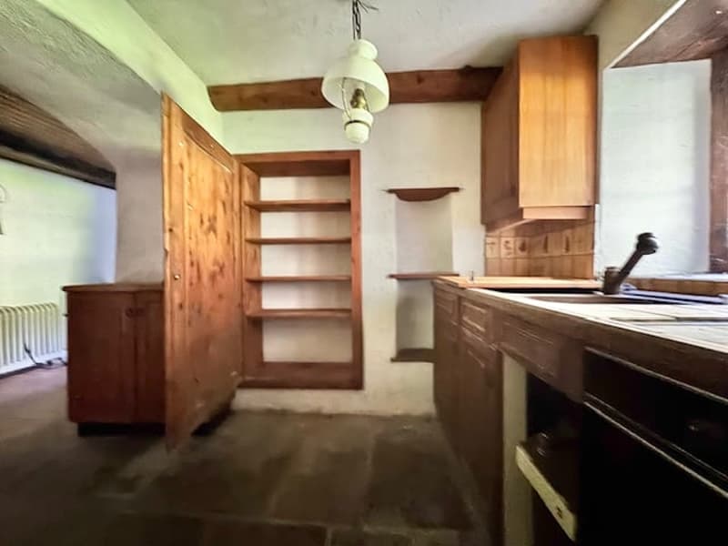 The image shows a rustic kitchen with wooden cabinets, shelves, and a wooden table or counter. The room has a stone or tile floor and exposed wooden beams on the ceiling.