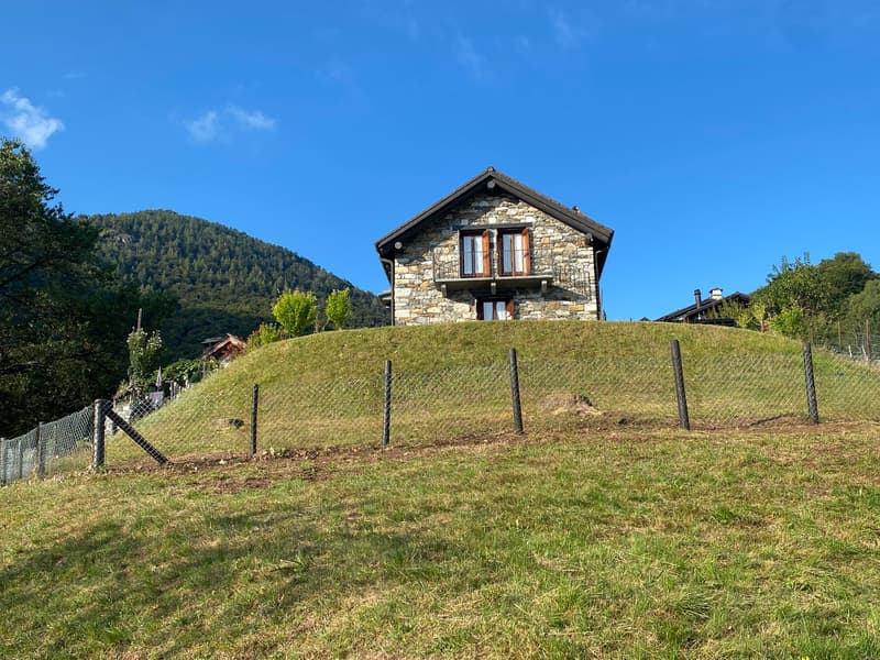 A stone house with a sloped roof is situated on top of a grassy hill with a fence, surrounded by trees and mountains