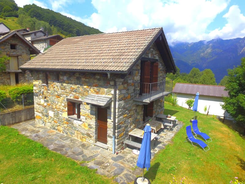 Two-story stone house with a tiled roof, surrounded by a grassy yard and mountains in the background. The house has a balcony or terrace, and there are blue patio umbrellas and lounge chairs visible.