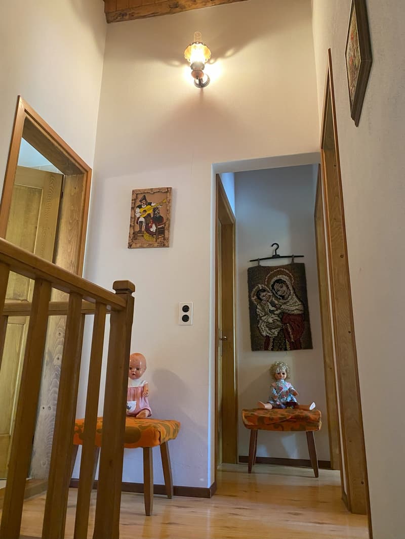 The image shows the interior of a room with wooden beams on the ceiling, a wooden staircase, and various decorative elements such as a framed artwork, a tapestry, and a wooden table with a doll on it.