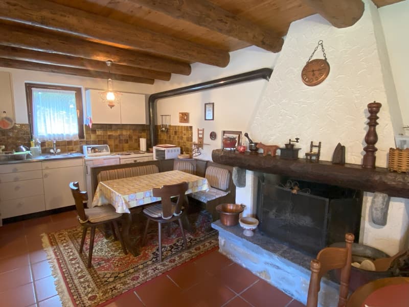 Rustic kitchen with wooden beams on the ceiling, tiled backsplash, dining table with chairs, and a fireplace with decorative items on the mantel.