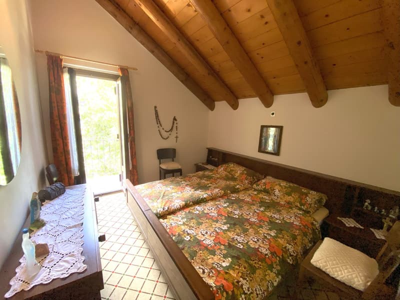 Bedroom with wooden ceiling beams, large window with access to a balcony or terrace, floral patterned bedding, wooden furniture, and decorative wall accents.