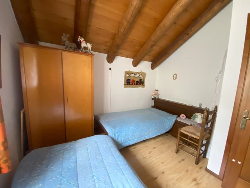 The image shows a cozy bedroom with a wooden ceiling and walls. There is a wooden wardrobe, two twin beds with blue bedding, and a small wooden chair. The room has a rustic, cabin-like feel to it.