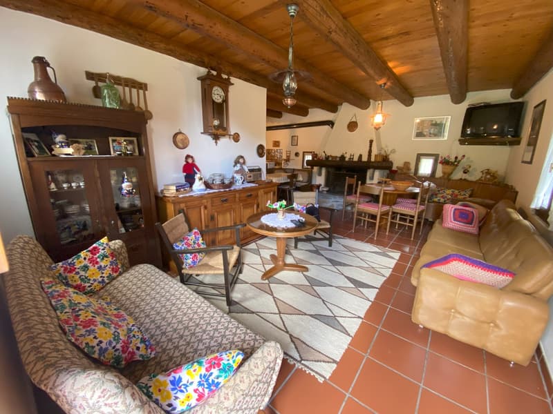Rustic interior with wooden beams, a fireplace, a large wooden cabinet, a round wooden table with chairs, and a couch with colorful floral patterns. The room has a mix of traditional and eclectic decor elements.