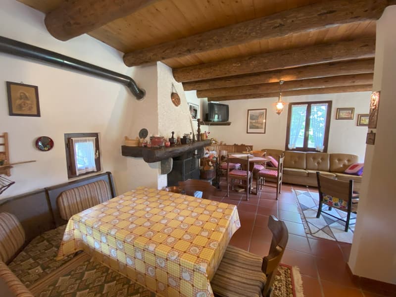 Rustic interior with exposed wooden beams, a fireplace, a dining table with a checkered tablecloth, and a sofa and chairs. The room has a cozy, traditional feel with various decorative items and artwork on the walls.