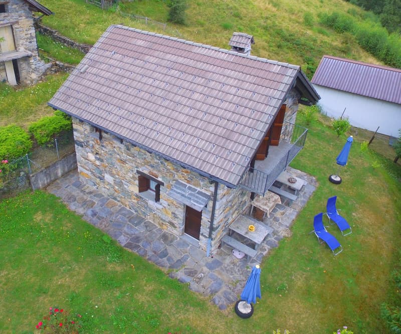 2-story stone house with a tiled roof, wooden balcony, garage, and parking space on a grassy hillside surrounded by greenery
