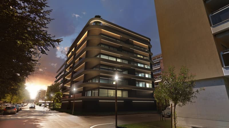 Modern residential building with glass and metal facade, multiple floors, illuminated at dusk, located in urban setting with street trees, parking and streetlights visible.