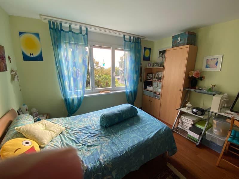 A bedroom with a bed, pillows, blue bed sheets, blue curtains, a wooden cabinet, shelves, and a window with a view outside